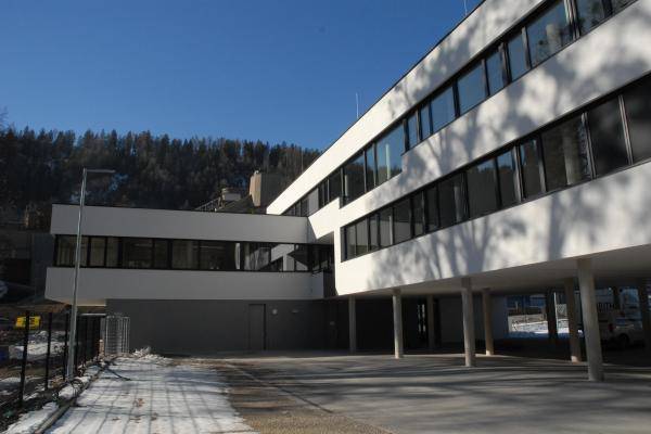 Aufnahme aus dem Hinterhof auf das Gebäude. Der linke Teil des L-Baus ist zwei Geschosse hoch, der rechte Teil drei. Im Erdgeschuss des rechten Gebäudeteils sind Stellplätze für PKW zwischen Betonsäulen. Zu sehen sind die weißen Fassaden des Gebäudes sowie die grau gerahmten Fenster, die auf beiden Stockwerken durchlaufend sind. Aufnahme aus dem Hinterhof auf das Gebäude. Der linke Teil des L-Baus ist zwei Geschosse hoch, der rechte Teil drei. Im Erdgeschuss des rechten Gebäudeteils sind Stellplätze für PKW zwischen Betonsäulen. Zu sehen sind die weißen Fassaden des Gebäudes sowie die grau gerahmten Fenster, die auf beiden Stockwerken durchlaufend sind.
