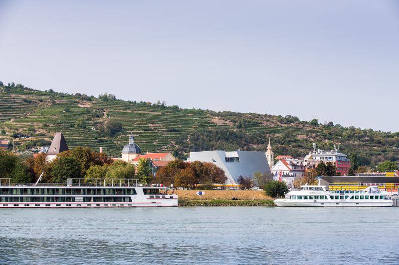 Im Vordergrund ist ein See zu sehen. Die Landesgalerie mit ihrer einzigartigen Form ist zwischen Bäumen hindurch in der Ferne zu sehen. Am Ufer des Sees sind zwei Schiffe zuz sehen. Im Hintergrund an einem Hang sind Weinreben zu erkennen. 
