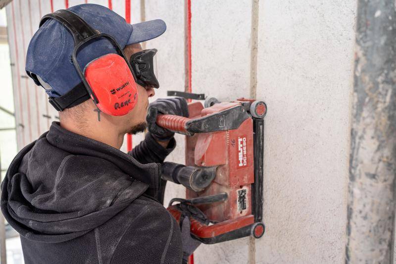 Ein Arbeiter mit rotem Gehörschutz und Schutzbrille fräst mit einer roten Maschine der Firma Hilti die Fugen für die Leitungen in die alte, verputzte Fassade. Im hintergrund sind bereits rote Leitungen in der Wand zu sehen.