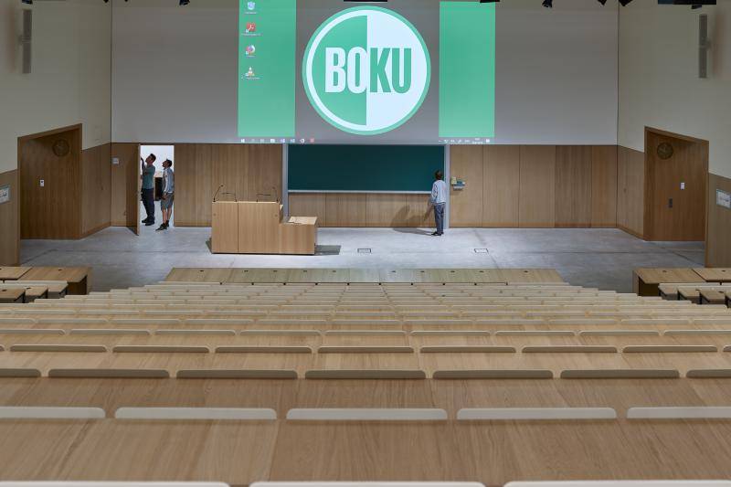 Blickvon der letzten Reihe im Hörsaal. Der Beamer projiziert das Logo der BOKU an die Wand. Die Tische sind ebenfalls mit Holzoberfläche, wie die Wandverkleidungen. Blickvon der letzten Reihe im Hörsaal. Der Beamer projiziert das Logo der BOKU an die Wand. Die Tische sind ebenfalls mit Holzoberfläche, wie die Wandverkleidungen.