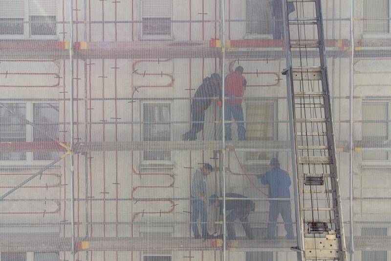 Hinter den Schutznetzen des Gerüsts sind Arbeiter zu sehen, die zwischen den Fenstern die roten Leitungen verlegen und besfestigen.