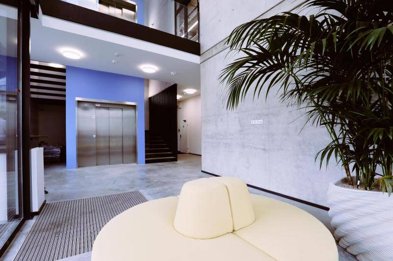 Innenaufnahmeaus dem Foyer. Sichtbetonwände, Aufzug, Treppenhaus und eine Sitzgelegenheit in gelb.