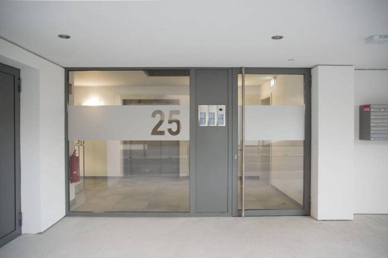 Eingang des Gebäudes. Große Glasfront und Glastüre mit grauem Rahmen und Mittelstück an dem die Klingeln angebracht sind. Eingang des Gebäudes. Große Glasfront und Glastüre mit grauem Rahmen und Mittelstück an dem die Klingeln angebracht sind.