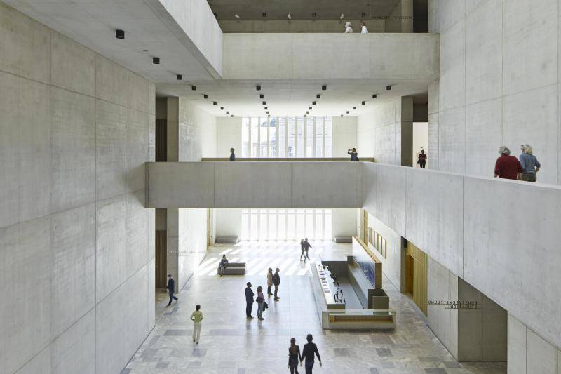 Das große und offene Foyer mit Blick von der Galerie aus dem ersten Stock. Wände und Geländer sind aus Sichtbeton. Die schwarzen Lampenan den Decken sind in einem Raster angeordnet. Vereinzelt sindMenschen alleine oder paarweise im Raum bzw. auf der Galeria zu sehen. Im Erdgeschoss des Foyers bickt man von obene auf einen Empfangstresen und eine kleine Sitzgelegenheit. Das große und offene Foyer mit Blick von der Galerie aus dem ersten Stock. Wände und Geländer sind aus Sichtbeton. Die schwarzen Lampenan den Decken sind in einem Raster angeordnet. Vereinzelt sindMenschen alleine oder paarweise im Raum bzw. auf der Galeria zu sehen. Im Erdgeschoss des Foyers bickt man von obene auf einen Empfangstresen und eine kleine Sitzgelegenheit.
