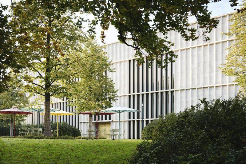 Im Vordergrund sind Büsche und Bäume zu sehen. Dazwischen stehen fünf Schirme in den Farben gelb, rot und türkis. Darunter stehen Stühle auf der Museumsterrasse. im Hintergrund ist dann die Fassade des Museums zu sehen mit der markanten Säulenoptik und großen Fenstern dahinter. Die Bäume bilden einen Rahmen um die Gebäudefassade aus dieser Bildperspektive. Im Vordergrund sind Büsche und Bäume zu sehen. Dazwischen stehen fünf Schirme in den Farben gelb, rot und türkis. Darunter stehen Stühle auf der Museumsterrasse. im Hintergrund ist dann die Fassade des Museums zu sehen mit der markanten Säulenoptik und großen Fenstern dahinter. Die Bäume bilden einen Rahmen um die Gebäudefassade aus dieser Bildperspektive.