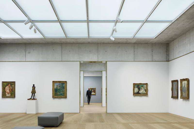 Zu sehen ist ein Ausstellungsraum des Museums. Die unteren dreiviertel der Wände sind weiß. Darauf hängen Gemälde in goldenen Rahmen. Das obere Viertel ist aus Beton. Die Decke besteht aus Milchglasscheiben, die für die Belichtung des Raumes sorgen. Der Boden besteht aus Holzdielen, auf denen zwei graue Sitzbänke stehen. Durch einen Durchgang in der Mitte des Bildes ist ein Mann zu sehen, der den Raum betritt. Zu sehen ist ein Ausstellungsraum des Museums. Die unteren dreiviertel der Wände sind weiß. Darauf hängen Gemälde in goldenen Rahmen. Das obere Viertel ist aus Beton. Die Decke besteht aus Milchglasscheiben, die für die Belichtung des Raumes sorgen. Der Boden besteht aus Holzdielen, auf denen zwei graue Sitzbänke stehen. Durch einen Durchgang in der Mitte des Bildes ist ein Mann zu sehen, der den Raum betritt.