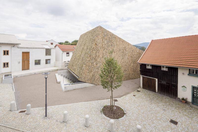 Der Würfel ragt aus demBoden. Umgeben vom Dorfplatz und der Bestandsbebauung. In der Mitte des Bildes ist ein frisch gepflanzert Baum zu sehen. Rechts neben dem Konzerthaus sieht man ein altes Bauernhaus. Der Würfel ragt aus demBoden. Umgeben vom Dorfplatz und der Bestandsbebauung. In der Mitte des Bildes ist ein frisch gepflanzert Baum zu sehen. Rechts neben dem Konzerthaus sieht man ein altes Bauernhaus.