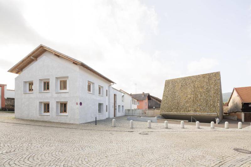 Ansicht auf den Platz mit Konzerthaus. Links ist ein weiterer Neubau aus Beton zu sehen, mit Holzfenstern und Dachüberstand mit Holzschalung. Rechts daneben ragt der Würfel des Konzerthauses aus dem Boden. Ansicht auf den Platz mit Konzerthaus. Links ist ein weiterer Neubau aus Beton zu sehen, mit Holzfenstern und Dachüberstand mit Holzschalung. Rechts daneben ragt der Würfel des Konzerthauses aus dem Boden.