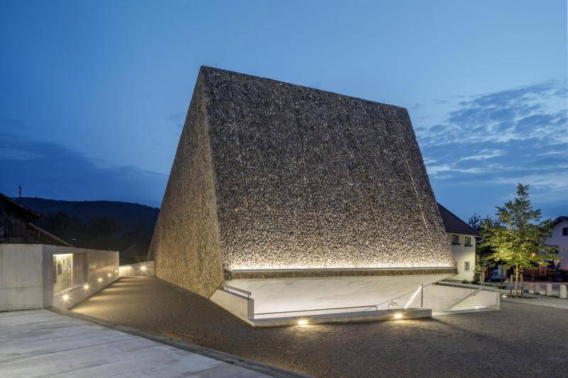 Aufnahme bei Dämmerung. Der Würfel aus braunen Steinen wird von unten beleuchtet. Auch der Eingang unter dem Würfel aus Sichtbeton ist hell erleuchtet. Aufnahme bei Dämmerung. Der Würfel aus braunen Steinen wird von unten beleuchtet. Auch der Eingang unter dem Würfel aus Sichtbeton ist hell erleuchtet.