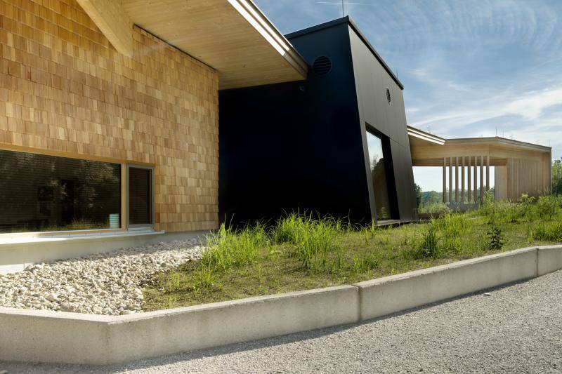 Ein Teil des Gebäudes ist mit schwarzen Fassadenplatten verkleidet. Der Rest des Gebäudes mit Holzschindeln. Vor dem Gebäude ist ein leicht erhöhtes Beet angelegt mit hohem Gras. Ein Teil des Gebäudes ist mit schwarzen Fassadenplatten verkleidet. Der Rest des Gebäudes mit Holzschindeln. Vor dem Gebäude ist ein leicht erhöhtes Beet angelegt mit hohem Gras.