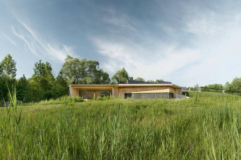 Das Gebäude fügt sich durch das Holz wunderbar in das Grün der Landschaft ein. Durch den eingeschossigen Bau verschwindet es sogar teilweise hinter den kleinen Erhebungen der Umgebung. Das Gebäude fügt sich durch das Holz wunderbar in das Grün der Landschaft ein. Durch den eingeschossigen Bau verschwindet es sogar teilweise hinter den kleinen Erhebungen der Umgebung.