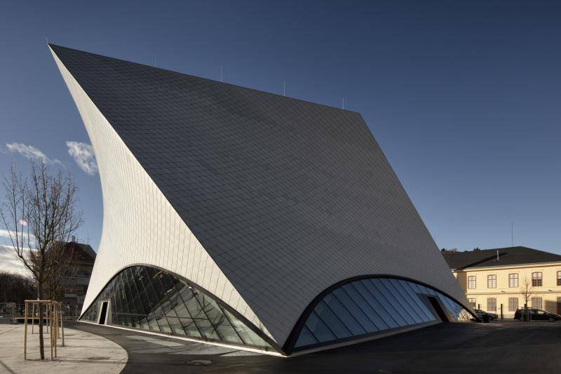 Die Schräggeneigte Fassade des Gebäudes ist in voller größe zu sehen. Unten ist ein Halbkreis aus Glas angeordnet. Die linke Seite des Gebäudes, die nach oben spitz zuläuft, ist von der Sonne angetrahlt.