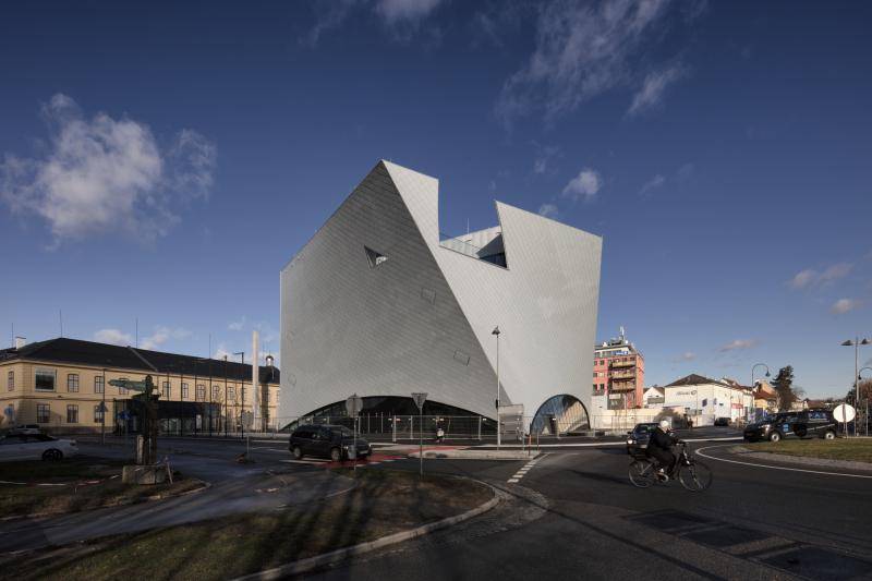 Das Gebäude ist hinter einer Straße zu sehen. Auf der Straße fahren Autos und ein Fahrrad. Oben am Gebäude ist ein Ausschnitt mit einem Glasgeländer zu sehen. Dort befindet sich eine Dachterrasse.