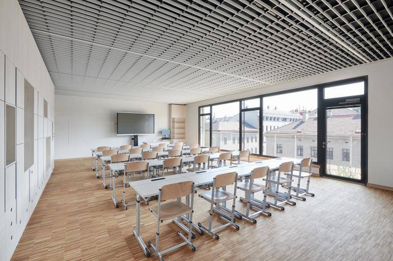 Innenaufnahme in einem Unterrichtsraum. Der Boden mit hellem Stäbchenparkett, weiße Wände, große Fensterfläche mit grauen Fensterrahmen. Tischreihen mit Stühlen in Richtung Smart-Board ausgerichtet. Decke mit weißen senkrecht stehenden Lamellen verkleidet.