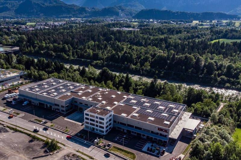 Blick aus der Luft auf die Halle. PV-Anlagen auf dem Flachdach zu sehen. Das Gebäude ist umgeben von Bäumen und eine kleinen Wald. Im weiten Hintergrund ist der Begin der Berge zu sehen. Blick aus der Luft auf die Halle. PV-Anlagen auf dem Flachdach zu sehen. Das Gebäude ist umgeben von Bäumen und eine kleinen Wald. Im weiten Hintergrund ist der Begin der Berge zu sehen.