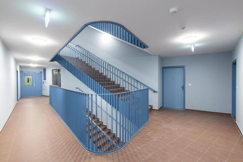 Eine Innenaufnahme aus dem Treppenhaus. Das Treppenauge hat eine Tropfenform. Die Wände sind in einem sehr hellen Blau und der Boden mit sandfarbenen Fliesen belegt. Das Geländer der Treppen ist blau und aus dünnen Querschnitten. Die Wohnungstüren sind ebenfalls blau, allerdings deutlich dunkler als die Wände und heben sich deutlich von diesen ab. Eine Innenaufnahme aus dem Treppenhaus. Das Treppenauge hat eine Tropfenform. Die Wände sind in einem sehr hellen Blau und der Boden mit sandfarbenen Fliesen belegt. Das Geländer der Treppen ist blau und aus dünnen Querschnitten. Die Wohnungstüren sind ebenfalls blau, allerdings deutlich dunkler als die Wände und heben sich deutlich von diesen ab.