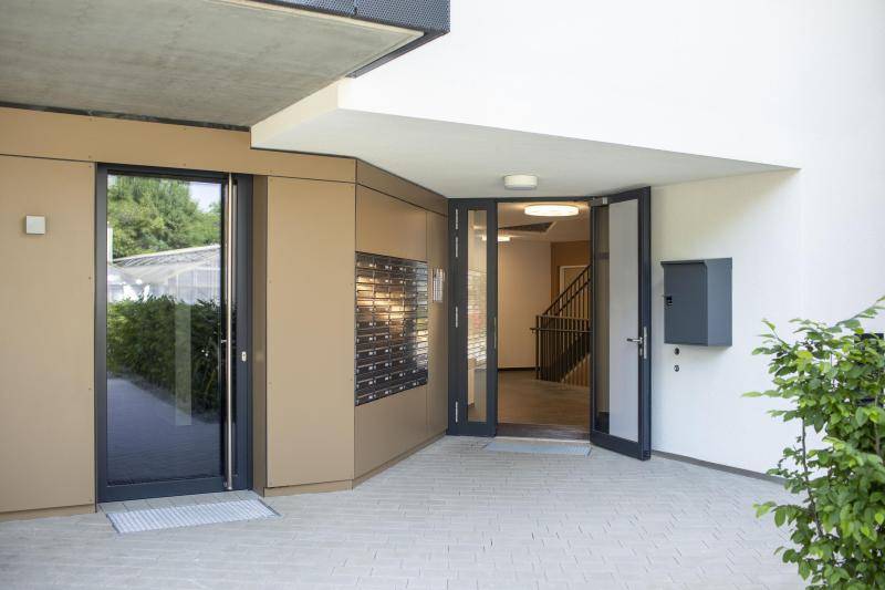 Der Eingang des Gebäudes mit großem Briefkasten in anthrazit in einer beigen Außenwand. Daneben die Haustüre, verglast. Der Eingang des Gebäudes mit großem Briefkasten in anthrazit in einer beigen Außenwand. Daneben die Haustüre, verglast.