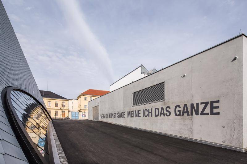Hier ist auf einer Sichtbetonfassade der Spruch "Wenn ich Kunst sage meine ich das Ganze" geschrieben. Links neben einem kleinen Hof steigt dann die Fassade der Galeria in die Höhe. Im Hintergrund sind andere Gebäude zu sehen.