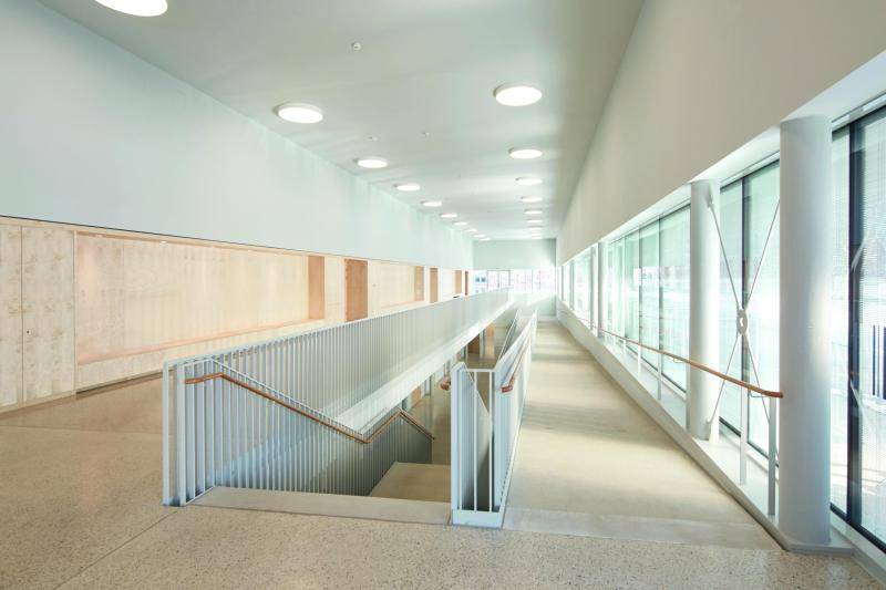Zu sehen, das Treppenhaus. in der Mitte läuft die Treppe nach unten ins Erdgeschoss. Rechts daneben erstreckt sich eine lange Rampe ins Erdgeschos. Rechts ist eine Fensterfront mit Säulen zu sehen. Links eine Wand, die in der unteren Hälfte aus Holz besteht und oben weiß verputzt ist. An der Decke sind runde Lichte angebracht. Das Tageslicht scheint durch die Fenster herrein Zu sehen, das Treppenhaus. in der Mitte läuft die Treppe nach unten ins Erdgeschoss. Rechts daneben erstreckt sich eine lange Rampe ins Erdgeschos. Rechts ist eine Fensterfront mit Säulen zu sehen. Links eine Wand, die in der unteren Hälfte aus Holz besteht und oben weiß verputzt ist. An der Decke sind runde Lichte angebracht. Das Tageslicht scheint durch die Fenster herrein