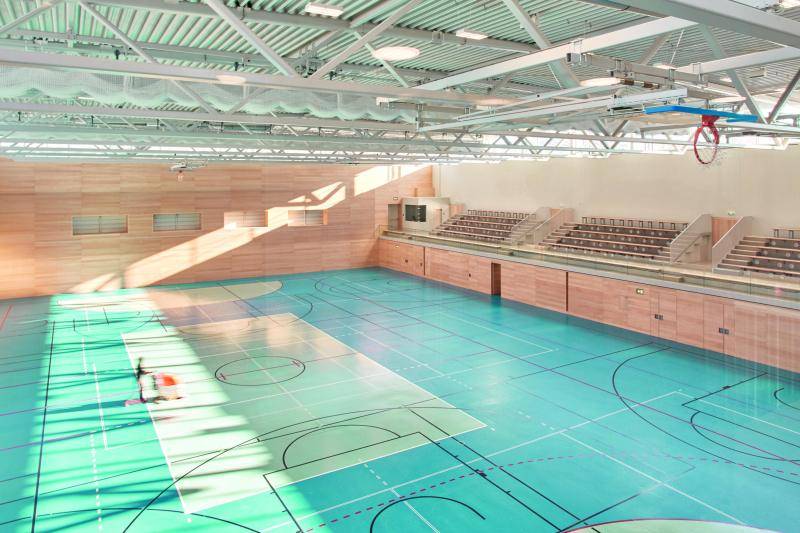 Der Blick von oben in die große Sporthalle. Die Wände aus Holz, ebenso die kleine Tribüne auf der rechten Seite. Die Dachkonstruktion sind weiße Fachwerkträger. Auf dem Hallenboden sind viele Linien für Spielfelder verschiedenster Sportarten eingezeichnet.  Der Blick von oben in die große Sporthalle. Die Wände aus Holz, ebenso die kleine Tribüne auf der rechten Seite. Die Dachkonstruktion sind weiße Fachwerkträger. Auf dem Hallenboden sind viele Linien für Spielfelder verschiedenster Sportarten eingezeichnet.