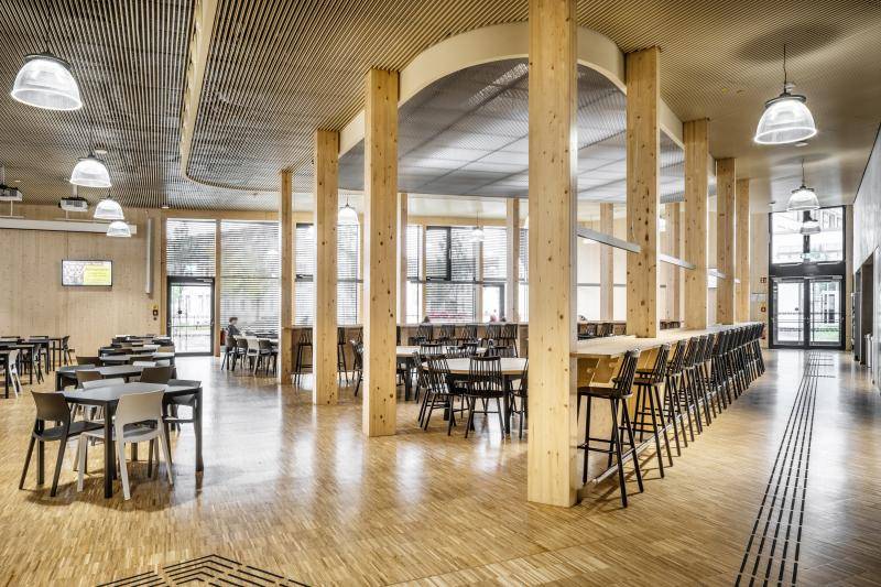 Innenraum der Mensa. Stäbchenparkett, Holzlamellen an der Decke, und Pfosten aus Brettschichtholz in der Raummitte. Verteilt im Raum sind Tische mit Stühlen zum Aufenthalt sowie ein länglicher Stehtisch mit Barhockern. Innenraum der Mensa. Stäbchenparkett, Holzlamellen an der Decke, und Pfosten aus Brettschichtholz in der Raummitte. Verteilt im Raum sind Tische mit Stühlen zum Aufenthalt sowie ein länglicher Stehtisch mit Barhockern.