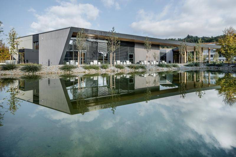 Das Gebäude mit grauer Fassade und den Fensterfronten spiegelt sich im davor liegenden See. Einige junge Bäume sind vor der Mensa gepflanz worden. Das Gebäude mit grauer Fassade und den Fensterfronten spiegelt sich im davor liegenden See. Einige junge Bäume sind vor der Mensa gepflanz worden.