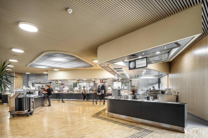 Essensausgabe. Dunkelgraue Selbstbedienungs Theken in geschwungener Linie angeordnet. Eine Theke für Salate etc. in der Raummitte. Sehr heller Raum durch Parkett, Holzwände und Holzlamellen an der Decke. Essensausgabe. Dunkelgraue Selbstbedienungs Theken in geschwungener Linie angeordnet. Eine Theke für Salate etc. in der Raummitte. Sehr heller Raum durch Parkett, Holzwände und Holzlamellen an der Decke.