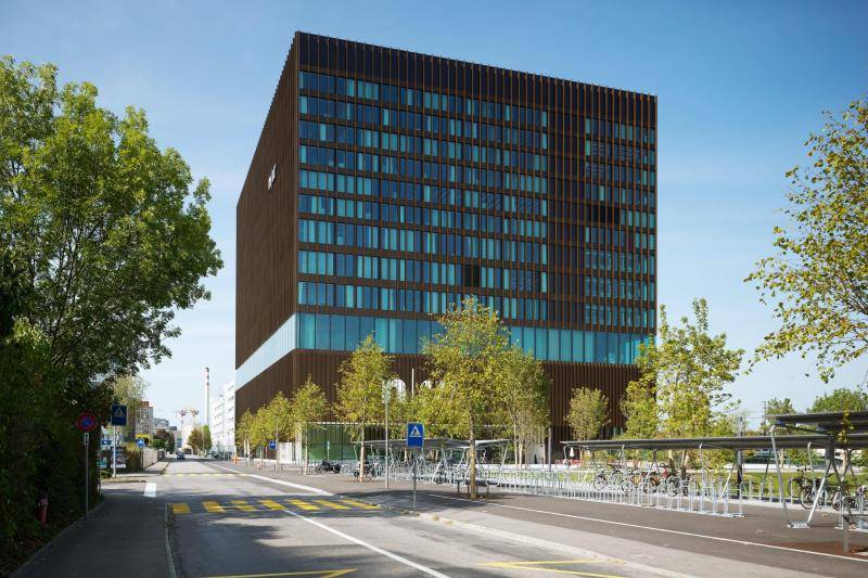 An der Fassade schimmern die großen Glasflächen türkis. Unterbrohen werden die Scheiben durch braune, metall Stäbe, die die Fassade gestalten. Das vierte Geschoss ist vollverglast, sodass die darüberliegenden Geschosse wie schwebend wirken.