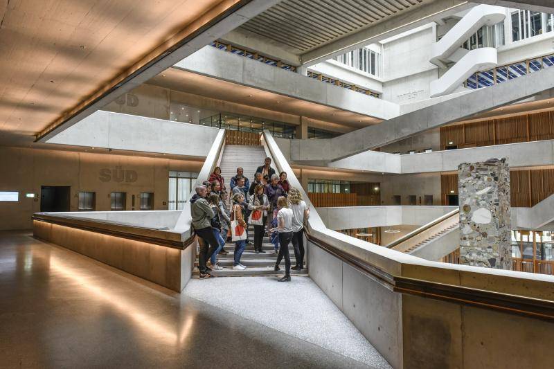 Eine Gruppe von Besucher*innen steht auf einer der Treppen im großen Foyerraum. Die Geländer sind aus Sichtbeton mit Holzhandläufen.