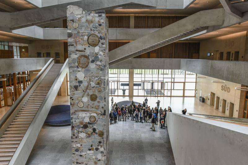 In der Mitte des großen Foyers, welches sich über die gesammte Gebäudehöhe öffnet, steht ein Betonpfeiler. Dieser ist gegossen mit Baumscheiben und großen Steinen an den Oberflächen.
