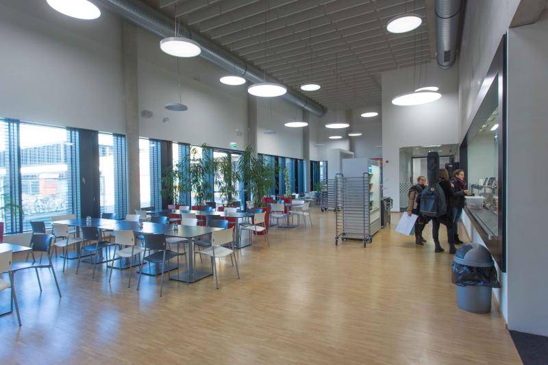 Dieses Bild zeigt das Innere der Mensa. Auf Holzfußboden sind einige Tische mit weißen und grauen Stühlen zu sehen. Drei Mädchen stehen an der Theke, die am rechten Bildrand zu sehen ist. Dieses Bild zeigt das Innere der Mensa. Auf Holzfußboden sind einige Tische mit weißen und grauen Stühlen zu sehen. Drei Mädchen stehen an der Theke, die am rechten Bildrand zu sehen ist.