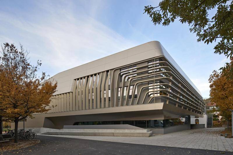 zu sehen ist ein Außeneck des Gebäudes. Hier ist die Fassade mit Aluminiumelementen verkleidet, die wie Stäbe erst senkrecht laufen und dann nach und nach um 90° in die Horizontale abknicken. Dahinter sind Fenster zu sehen, die dadurch je nach Blickwinkel verschattet werden. Über dem Gebäude ist der hellblaue Himmel zu sehen und am Bildrand stehen Bäume. zu sehen ist ein Außeneck des Gebäudes. Hier ist die Fassade mit Aluminiumelementen verkleidet, die wie Stäbe erst senkrecht laufen und dann nach und nach um 90° in die Horizontale abknicken. Dahinter sind Fenster zu sehen, die dadurch je nach Blickwinkel verschattet werden. Über dem Gebäude ist der hellblaue Himmel zu sehen und am Bildrand stehen Bäume.