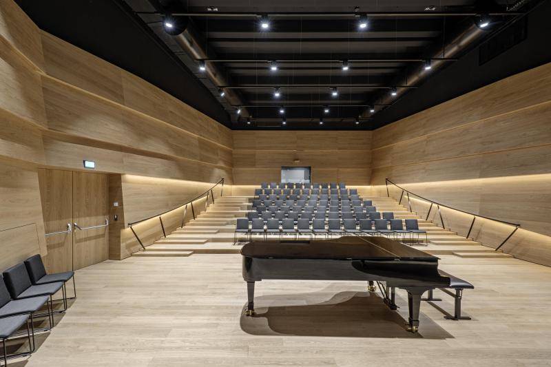 Zu sehen ist der Konzertraum des Gebäudes. Dieser ist mit hellen Holzoberflächen ausgekleidet. Auch der Boden ist aus Holz. Hinten stehen ca. 80 graue Stühle für Zuschauer*innen. Die Reihen sind wie eine Tribüne aufsteigend. Vorn auf der Bühne, die sich auf der Bodenebene befindet, steht ein schwarzer Flügel. Zu sehen ist der Konzertraum des Gebäudes. Dieser ist mit hellen Holzoberflächen ausgekleidet. Auch der Boden ist aus Holz. Hinten stehen ca. 80 graue Stühle für Zuschauer*innen. Die Reihen sind wie eine Tribüne aufsteigend. Vorn auf der Bühne, die sich auf der Bodenebene befindet, steht ein schwarzer Flügel.