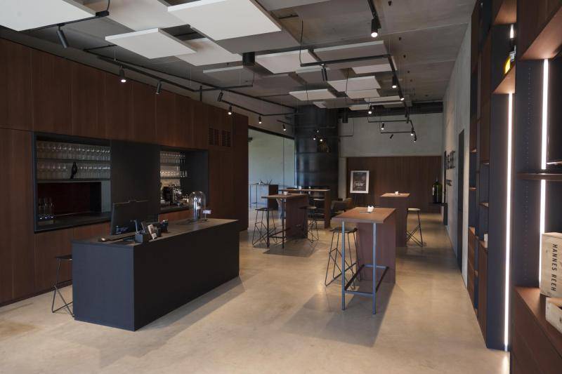 Ein Innenraum mit einer Art Bar, auf der ein Bildschirm steht. Dahinter eine Schrankwand mit vielen Weingläsern. Die verkleidung/ die Möbel des Raums sind mit dunklen Holzoberflächen. Im Raum stehen mehrere hohe Tische mit Bar-Stühlen. Ein Innenraum mit einer Art Bar, auf der ein Bildschirm steht. Dahinter eine Schrankwand mit vielen Weingläsern. Die verkleidung/ die Möbel des Raums sind mit dunklen Holzoberflächen. Im Raum stehen mehrere hohe Tische mit Bar-Stühlen.