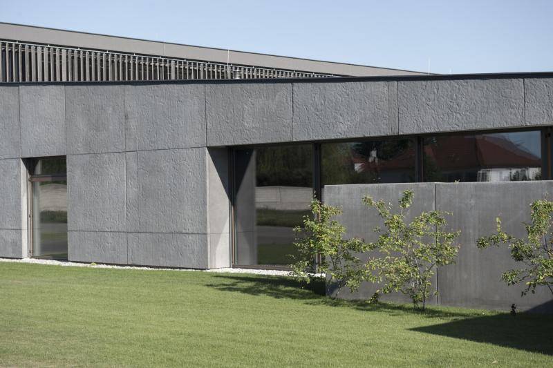 Die seitliche Fassade des Gebäudes aus großen, dunkel grauen Steinplatten. Darin Bodentiefe Fenster mit schwarzen Rahmen Die seitliche Fassade des Gebäudes aus großen, dunkel grauen Steinplatten. Darin Bodentiefe Fenster mit schwarzen Rahmen