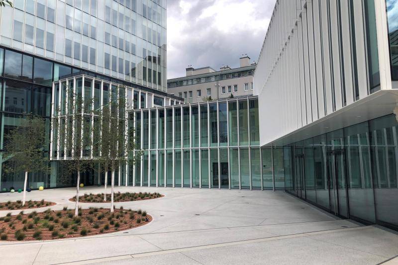 Der Vorplatz des Gebäudes mit seinen Hellen Betonplatten und den Beeten. Rechts im Bild der Gebäudeteil mit zwei Geschossen. Links beginnt der Hochhausteil des Komplexes. 