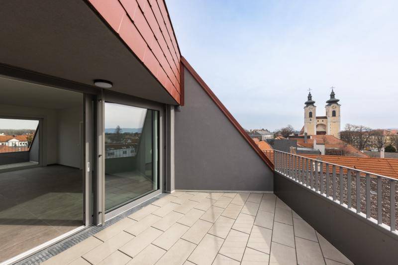 Zu sehen ist eine Dachloggia mit hellen Fliesen belegt. Die Wohnung ist durch eine große Schiebetüre geöffnet. Man blickt von der Terrasse über die Dächer der Stadt. Zu sehen ist eine Dachloggia mit hellen Fliesen belegt. Die Wohnung ist durch eine große Schiebetüre geöffnet. Man blickt von der Terrasse über die Dächer der Stadt.