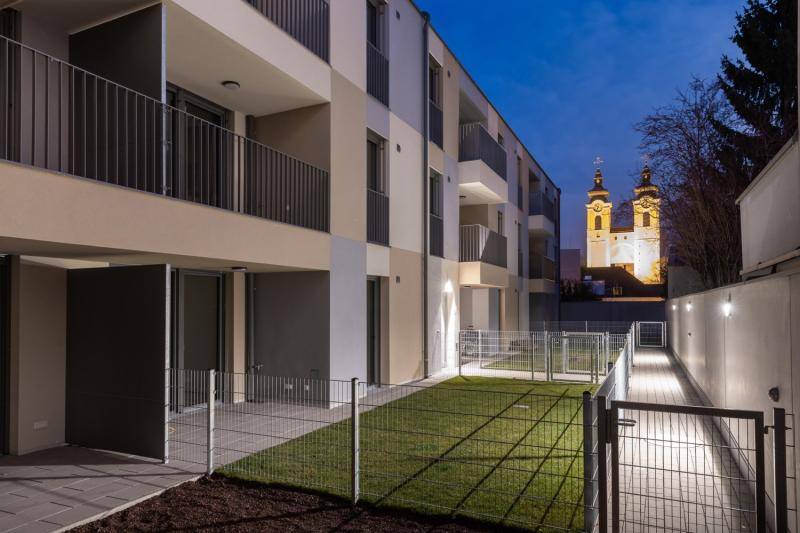 Im Erdgeschoss haben die Wohnungen kleine Terrassen, sowie Rasenflächen. Diesesindmit Zäunen eingegrenzt. Der daneben liegende Weg ist in der Dämmerung beleuchtet. Im Hintergrund idt die Kirche der Stadt hell erleuchtet. Im Erdgeschoss haben die Wohnungen kleine Terrassen, sowie Rasenflächen. Diesesindmit Zäunen eingegrenzt. Der daneben liegende Weg ist in der Dämmerung beleuchtet. Im Hintergrund idt die Kirche der Stadt hell erleuchtet.