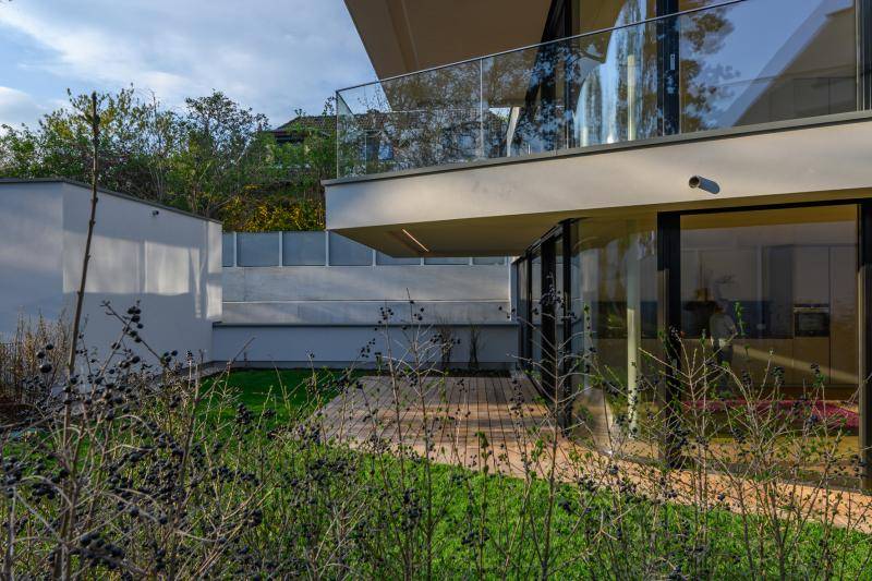 Das Gebäude mit seinen Balkonen im Hintergund. Im Vordergrund sind Sträucher zu sehen und der Rasen im Garten
