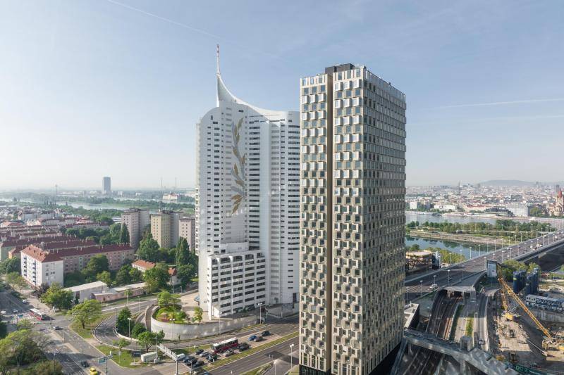 Luftaufnahme des Gebäudes. Im Hintergrund ist ein weiteres weißes Hochhaus zu sehen. Der DC Tower 3 hat eine einzigartige Fassade. Die Fenster stehen wie kleine Erker schräg aus der Fassade heraus. In jedem Stockwerk wechselt die Ausrichtung zwischen Links und Rechts. Im Hintergrund ist die Donau zu sehen. Das Gebäude ist umgeben von Straßen. Luftaufnahme des Gebäudes. Im Hintergrund ist ein weiteres weißes Hochhaus zu sehen. Der DC Tower 3 hat eine einzigartige Fassade. Die Fenster stehen wie kleine Erker schräg aus der Fassade heraus. In jedem Stockwerk wechselt die Ausrichtung zwischen Links und Rechts. Im Hintergrund ist die Donau zu sehen. Das Gebäude ist umgeben von Straßen.