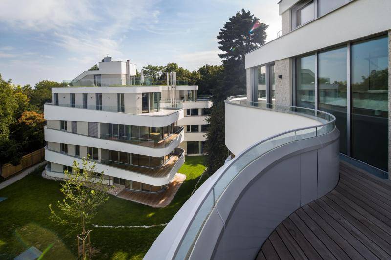 Die Aufnahme ist aus dem dritten Stock eines der Gebäude gemacht. Zu sehen ist an der Seite eine Holzterrasse und ein weises, massives Geländer. Unten ist grüne Wiese und ein frisch gepflanzter Baum zu sehen. Dahinter ist ein weiteres neues Gebäude zu sehen mit seinen geschwungenen, abgerundeten Ecken. Das weiße Geländer geht hier in Glas über und auch hier sind Terrassen/Balkone mit Holzbelag zu sehen. Die Aufnahme ist aus dem dritten Stock eines der Gebäude gemacht. Zu sehen ist an der Seite eine Holzterrasse und ein weises, massives Geländer. Unten ist grüne Wiese und ein frisch gepflanzter Baum zu sehen. Dahinter ist ein weiteres neues Gebäude zu sehen mit seinen geschwungenen, abgerundeten Ecken. Das weiße Geländer geht hier in Glas über und auch hier sind Terrassen/Balkone mit Holzbelag zu sehen.