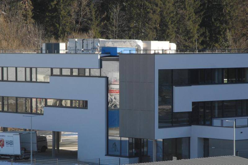 Aus der Distanz ist das gesamte Gebäude zu sehen. Der L-Förmige Bau ist weiß verputzt. Das Gebäude hat drei Stockwerke. Im linken Teil ist das unterste Geschoss eine Parkfläche für PKW. Im zweiten und dritten Geschoss sind Fenster aneinander gereiht mit grauen Rahmen. So sieht der Rest der weißen Fassade aus wie ein großes "S". Der rechte Teil der Gebäudes hat im 2. Obergeschoss zur Hälfte ein Flachdach und ist nicht vollständig bebaut. Auf dem Flachdach ist eine Lüftungsanlage zu sehen. Aus der Distanz ist das gesamte Gebäude zu sehen. Der L-Förmige Bau ist weiß verputzt. Das Gebäude hat drei Stockwerke. Im linken Teil ist das unterste Geschoss eine Parkfläche für PKW. Im zweiten und dritten Geschoss sind Fenster aneinander gereiht mit grauen Rahmen. So sieht der Rest der weißen Fassade aus wie ein großes "S". Der rechte Teil der Gebäudes hat im 2. Obergeschoss zur Hälfte ein Flachdach und ist nicht vollständig bebaut. Auf dem Flachdach ist eine Lüftungsanlage zu sehen.