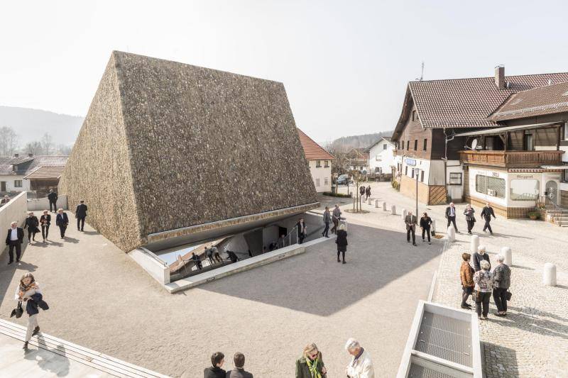 Das Konzerthaus ist geformt wie ein Würfel, der zur Hälfte in der Erde verschwindet. Allerdings würde der Würfel auf einer Kante stehen, also um 45° geneigt sein. Unter dem Würfel führen Treppen unter die Erde, wo sich der Eingang befindet. Der Dorfplatz in unmittelbarer Nähe darum ist mit einem Sand-Estrick bedeckt. Außenherum ist der Bereich gepflastert. Es sind viele Menschen über den Platz verteilt zu sehen. Das Konzerthaus ist geformt wie ein Würfel, der zur Hälfte in der Erde verschwindet. Allerdings würde der Würfel auf einer Kante stehen, also um 45° geneigt sein. Unter dem Würfel führen Treppen unter die Erde, wo sich der Eingang befindet. Der Dorfplatz in unmittelbarer Nähe darum ist mit einem Sand-Estrick bedeckt. Außenherum ist der Bereich gepflastert. Es sind viele Menschen über den Platz verteilt zu sehen.