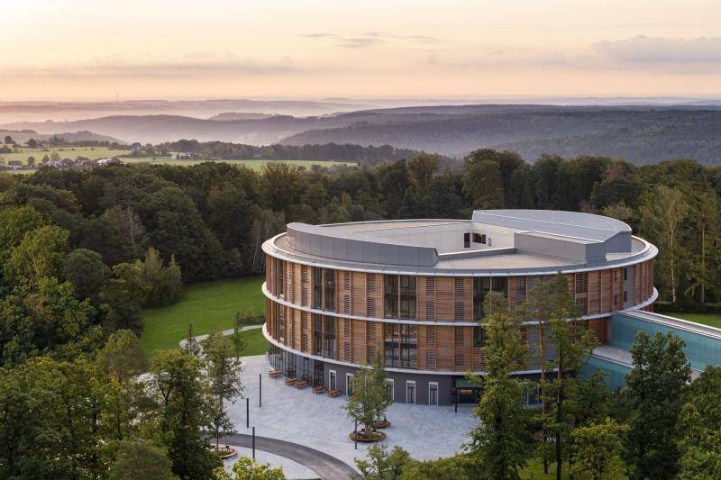Das runde Gebäude mit seiner Holz-Fassade in Mitten von Wald. Im Hintergrund ist der Sonnenuntergang zu sehen. Das runde Gebäude mit seiner Holz-Fassade in Mitten von Wald. Im Hintergrund ist der Sonnenuntergang zu sehen.