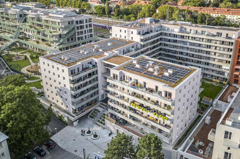 Blick von Oben auf das Gebäude. Das Gebäude ist ein L-Förmiger Bau mit einem kleineren Rechteckigen gebäude im Innenhof. Dieser ist durch ein Überdachtes Treppenhaus verbunden. Es gibt auf der gesamten Länge Balkone. Das Flachdach ist begrünt und mit PV-Anlagen belegt. Hinter dem Gebäude laufen die Bahngleise zum Hauptbahnhof. Blick von Oben auf das Gebäude. Das Gebäude ist ein L-Förmiger Bau mit einem kleineren Rechteckigen gebäude im Innenhof. Dieser ist durch ein Überdachtes Treppenhaus verbunden. Es gibt auf der gesamten Länge Balkone. Das Flachdach ist begrünt und mit PV-Anlagen belegt. Hinter dem Gebäude laufen die Bahngleise zum Hauptbahnhof.