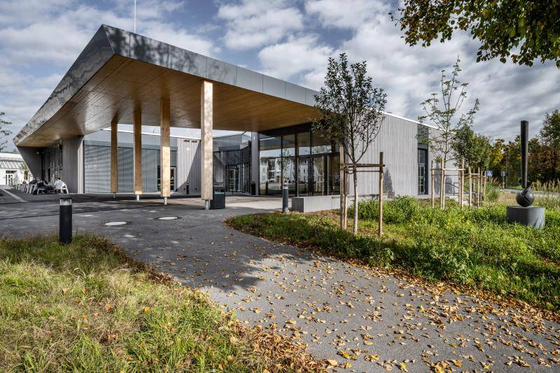 Eingang der Mensa mit Überdachtem Vorplatz. Die Holzkonstruktion ist durch Pfosten und Schalung an der Decke zu erkennen. Außen ist das Gebäude grau Verschalt. Tische mit Bänken zum Essen stehen vor der Glasfront. Eingang der Mensa mit Überdachtem Vorplatz. Die Holzkonstruktion ist durch Pfosten und Schalung an der Decke zu erkennen. Außen ist das Gebäude grau Verschalt. Tische mit Bänken zum Essen stehen vor der Glasfront.
