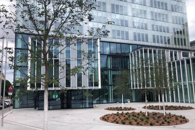 Vor dem Gebäude ist der Platz mit großen Betonplatten bedeckt, dazwischen sind runde Beete mit vielen kleinen Sträuchern und vereinzelt frisch gesetzten Bäumen. Dahinter steigt die Glasfassade des Gebäudes in die Höhe. Die Fassade besteht zur Gänze aus Glas, mit dünnen Pfosten in schwarz und weiß dazwischen. Vor dem Gebäude ist der Platz mit großen Betonplatten bedeckt, dazwischen sind runde Beete mit vielen kleinen Sträuchern und vereinzelt frisch gesetzten Bäumen. Dahinter steigt die Glasfassade des Gebäudes in die Höhe. Die Fassade besteht zur Gänze aus Glas, mit dünnen Pfosten in schwarz und weiß dazwischen.